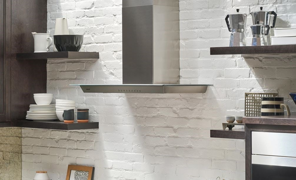 The Wolf 36&quot; Cooktop Wall Hood - Glass (VW36G) shown set against white exposed brick and floating shelves in a luxury kitchen design