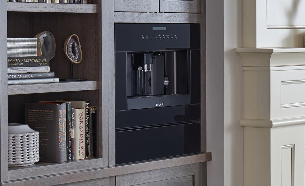 The Wolf 24&quot; Coffee System Black Glass (EC24/B) shown set seamlessly in bookshelf featuring warm wood tones and modern handles